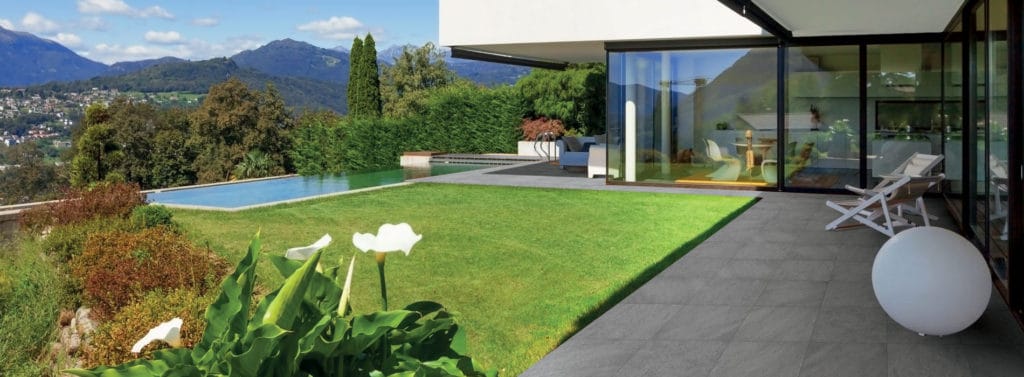 Esterno di una casa con giardino, pavimenti in gres Altra Pietra e piscina. Montagne si vedono in lontananza e dei fiori vicino all'osservatore